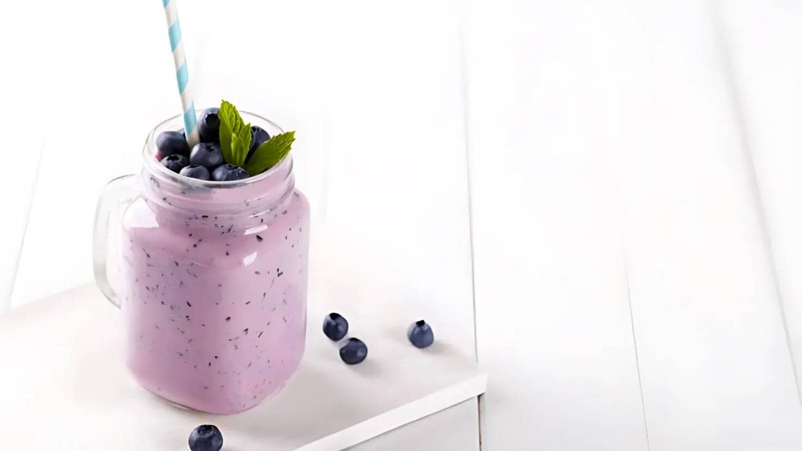 A mason jar filled with a purple blueberry smoothie, topped with blueberries and mint leaves, with a striped straw and scattered blueberries on a white surface.