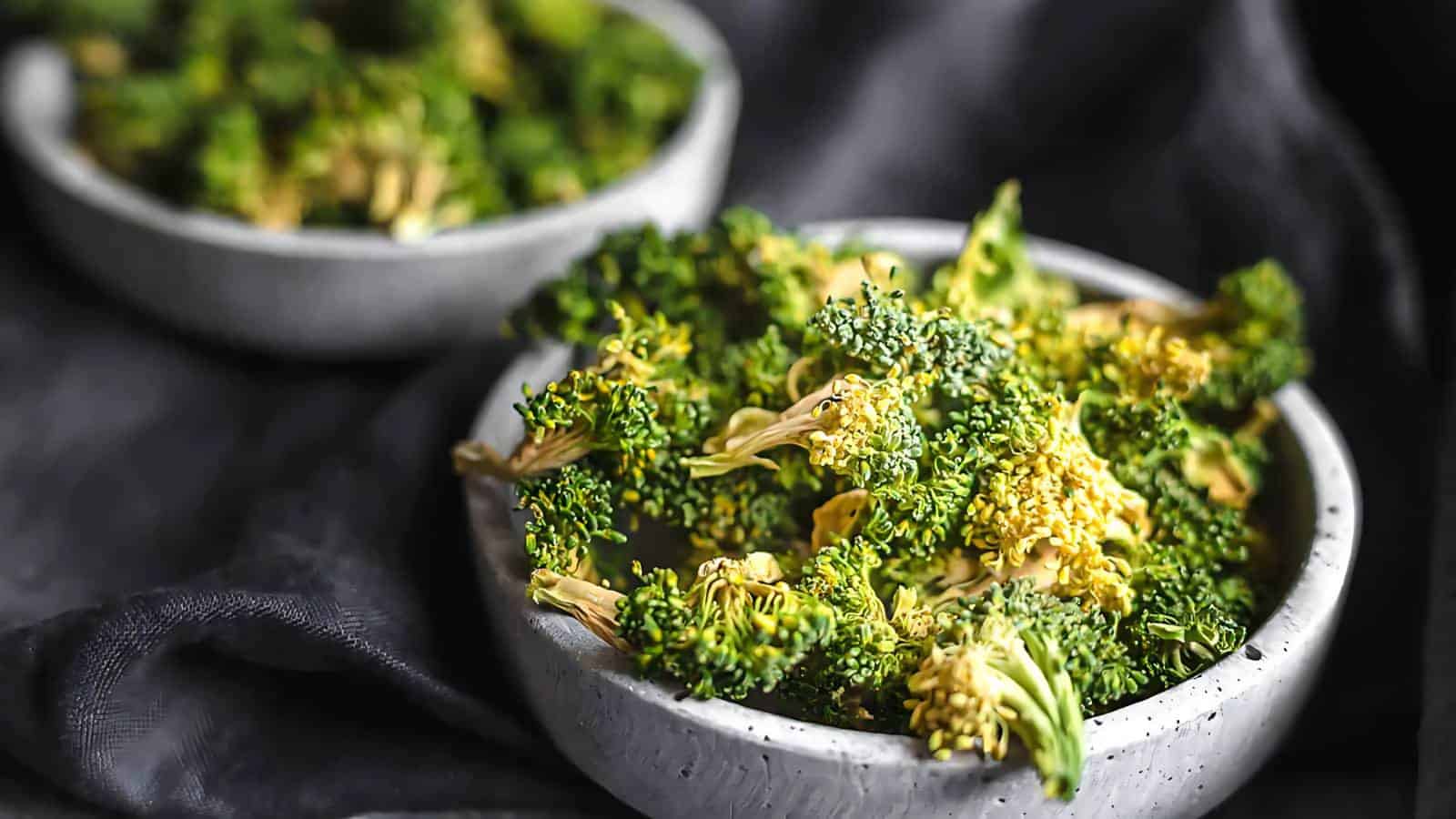 A bowl of fresh broccoli florets with another bowl of broccoli in the background, both placed on a dark surface.