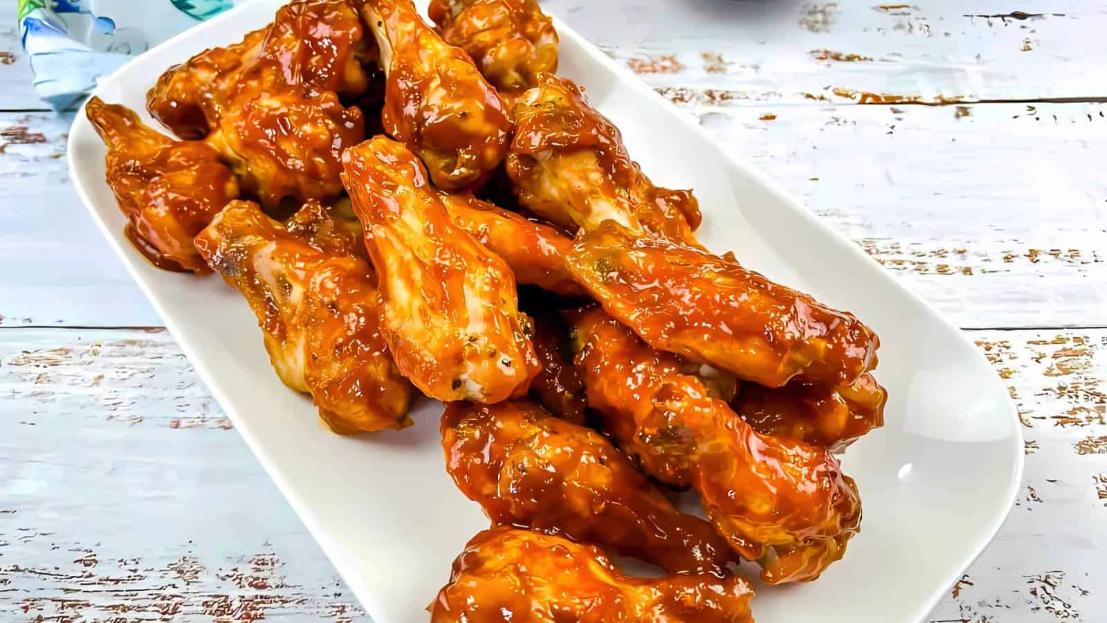 A white rectangular plate filled with glazed chicken wings coated in a reddish-brown sauce, placed on a rustic white wooden surface.