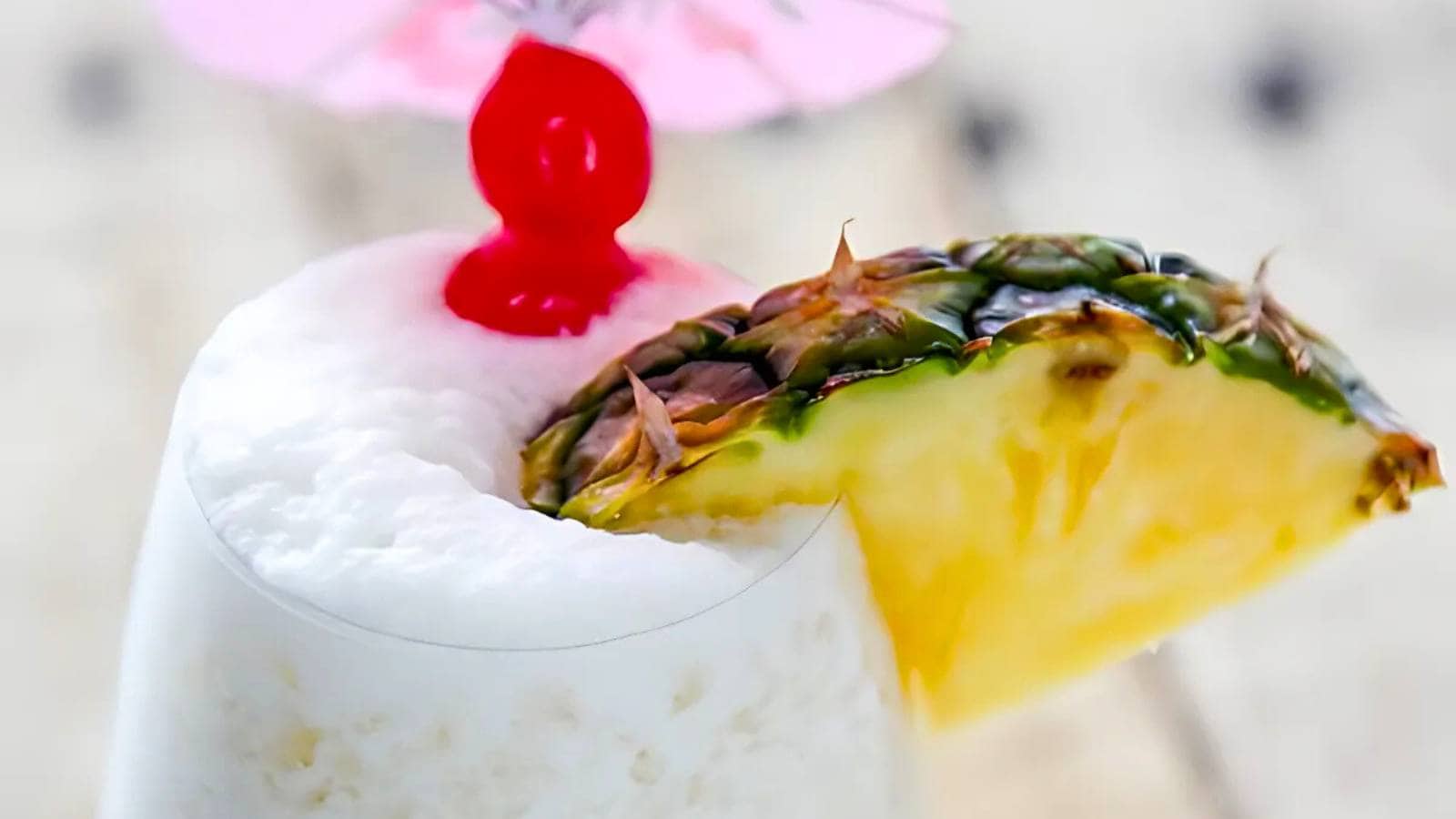 A glass of piña colada topped with a maraschino cherry, a slice of pineapple, and a pink cocktail umbrella.