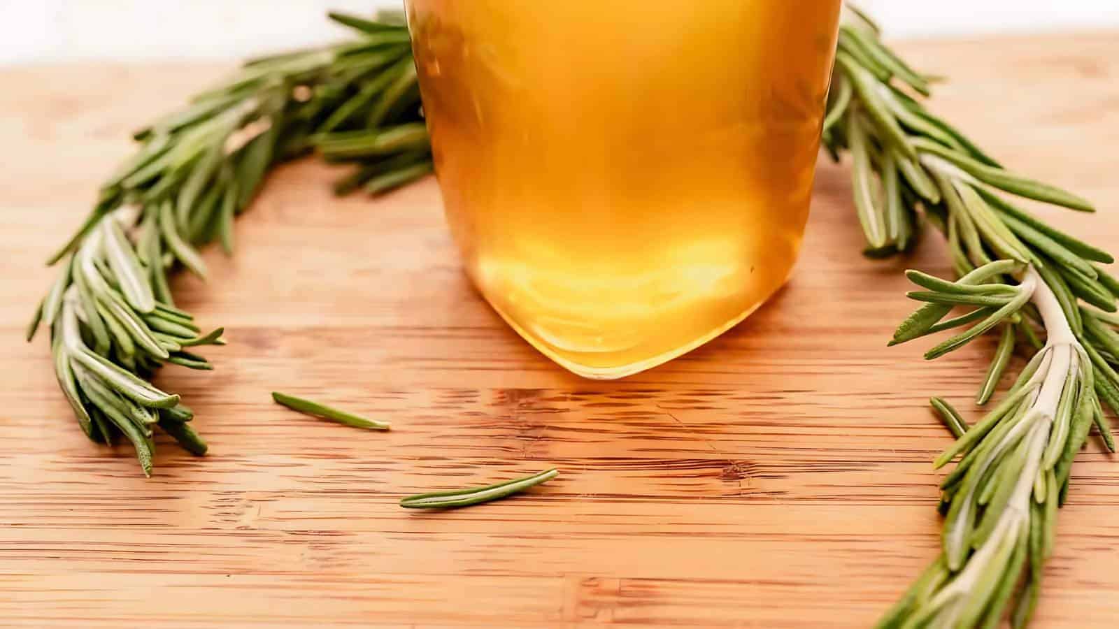 A glass jar filled with golden liquid sits on a wooden surface, surrounded by a curved sprig of fresh rosemary.