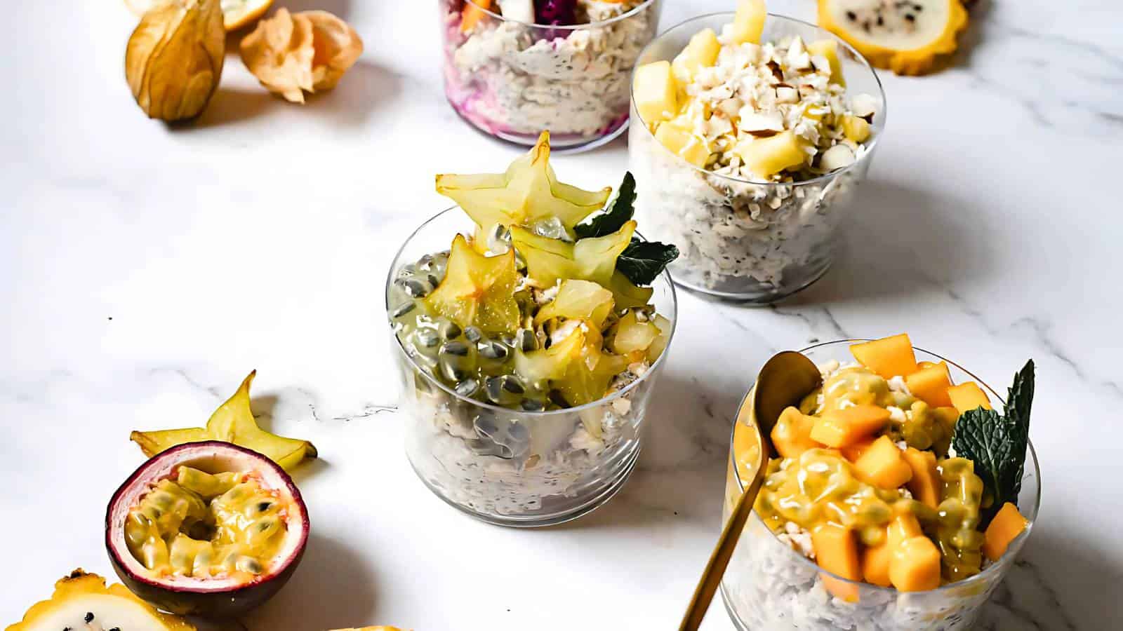 Four glass cups filled with oatmeal, topped with various fruits such as starfruit, passionfruit, melon, and shredded coconut, are arranged on a white marble surface.