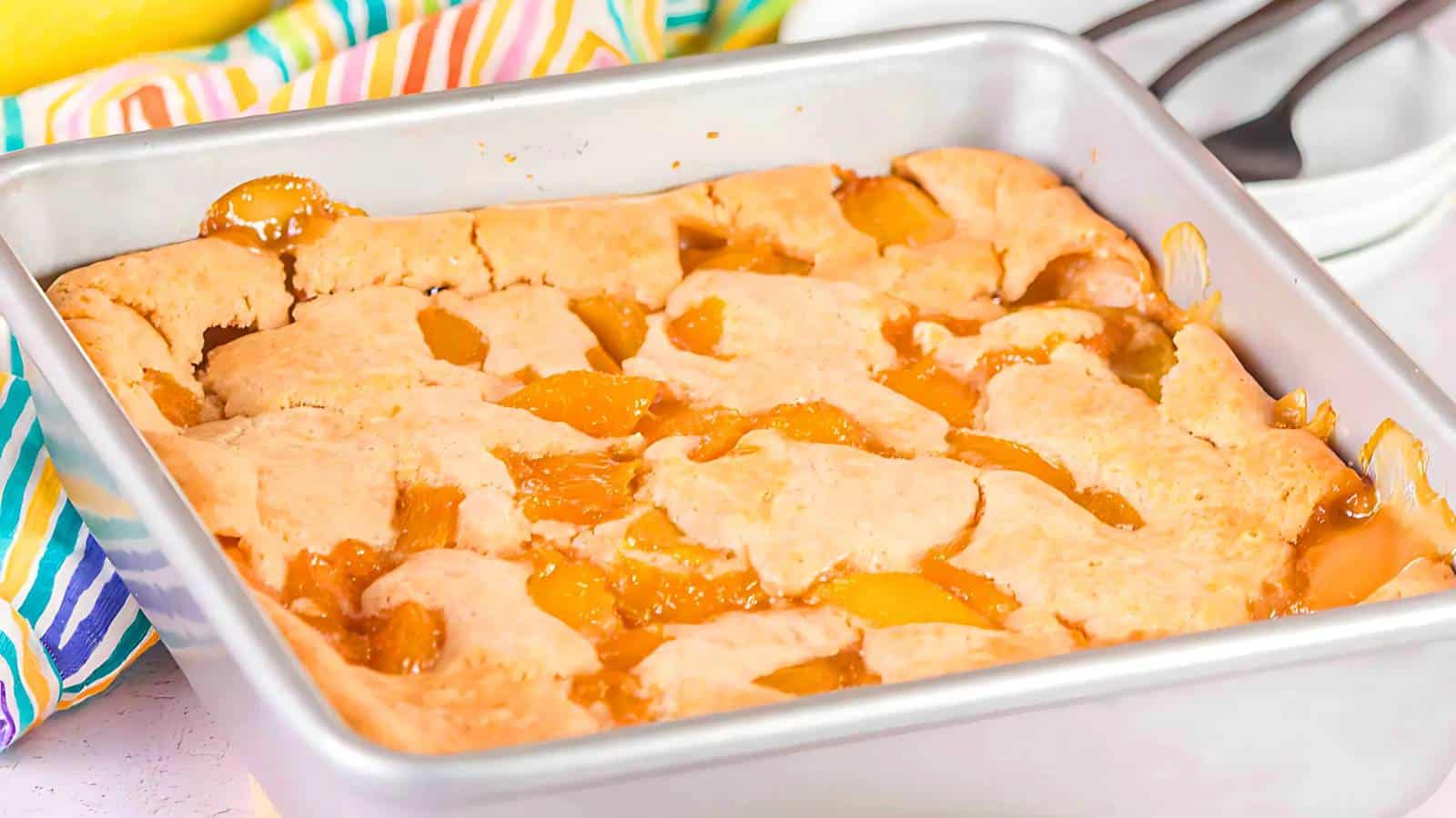 A square baking pan filled with golden-brown peach cobbler, featuring a crust with visible peach filling peeking through cracks.