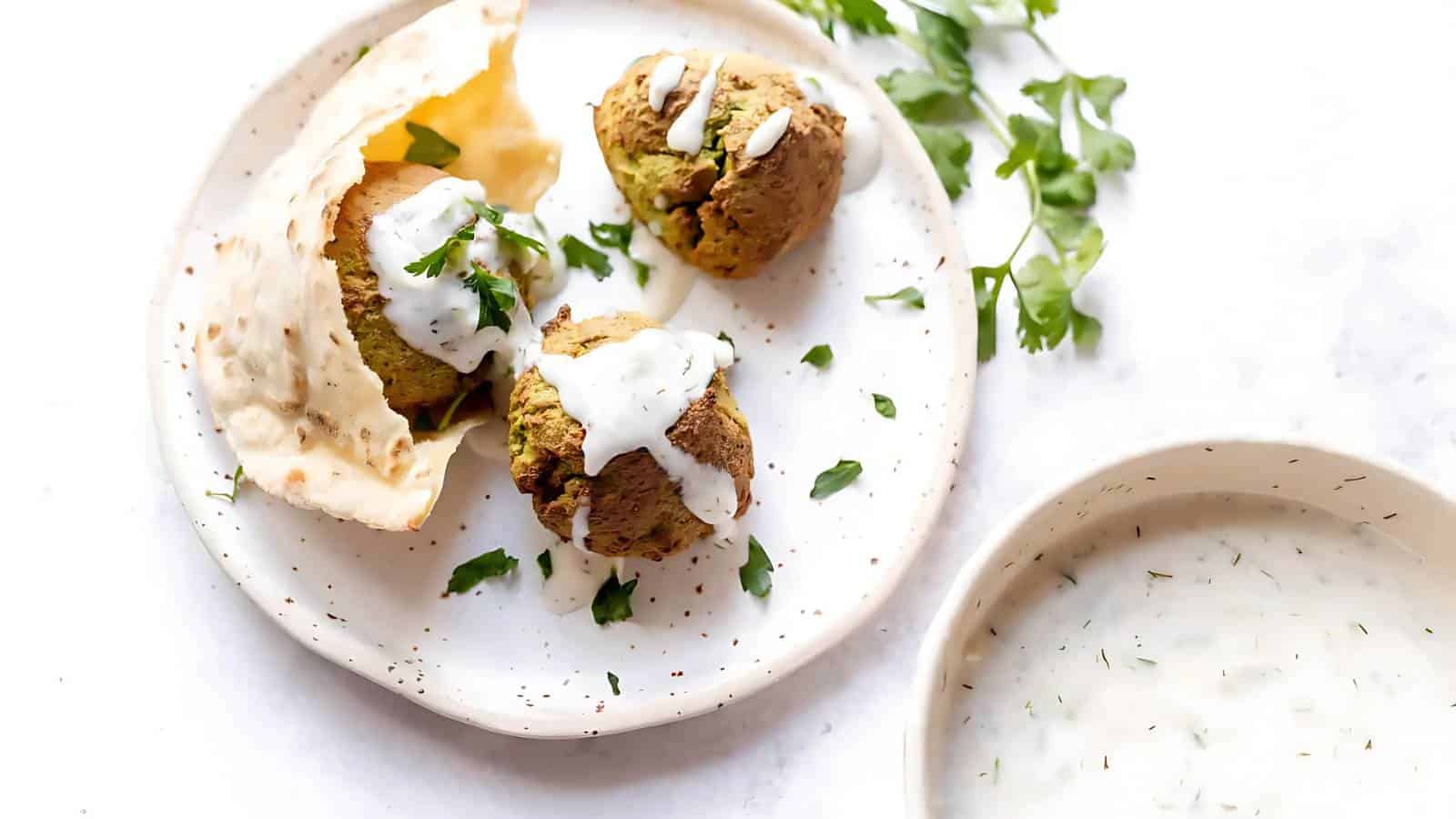 Three falafel balls with white sauce and herbs on a plate with flatbread, alongside a bowl of white sauce on a white background.