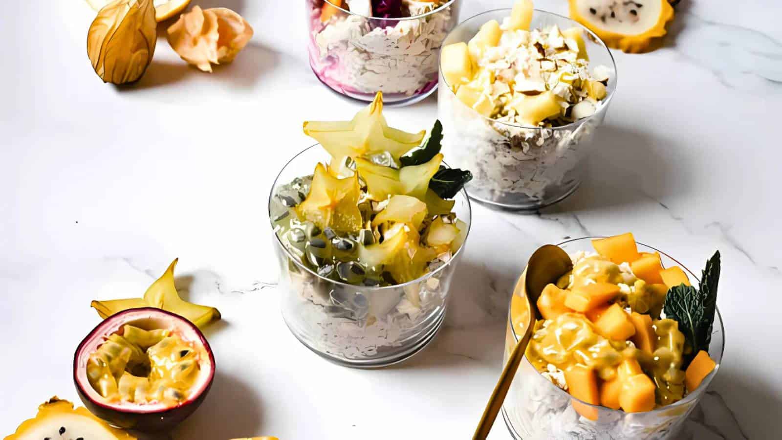 Four clear cups filled with yogurt, granola, and assorted fresh fruits including starfruit, passion fruit, melon, and physalis, are arranged on a white surface with a gold spoon.