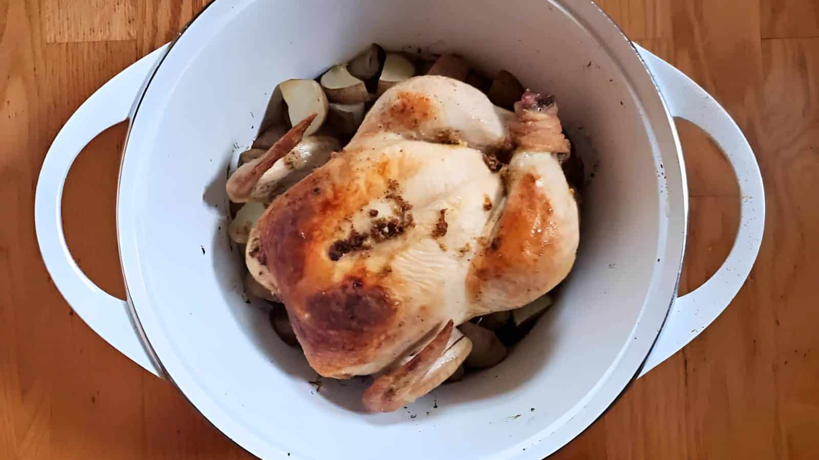 A whole roasted chicken with browned skin sits in a white Dutch oven on top of chopped potatoes.
