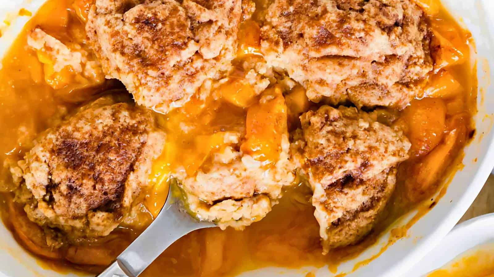 Close-up of peach cobbler with golden-brown biscuit topping and a spoon scooping out a portion.