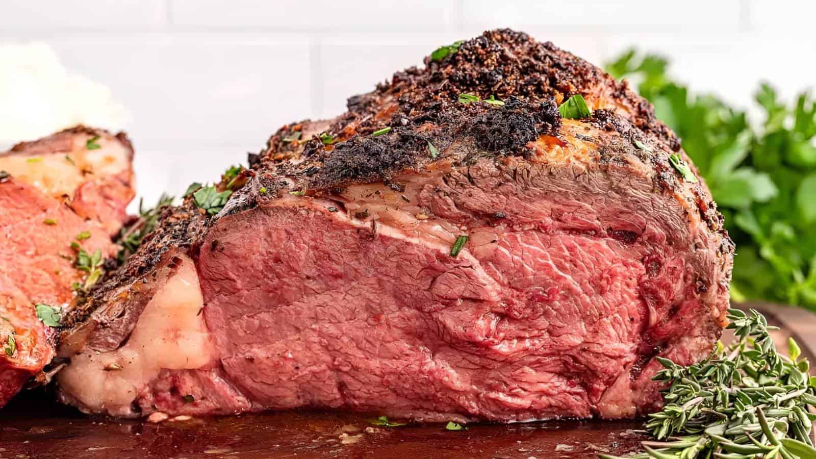 A cooked prime rib roast with a seasoned crust, sliced to reveal a juicy, medium-rare interior. Fresh herbs are visible next to the meat on a wooden surface.
