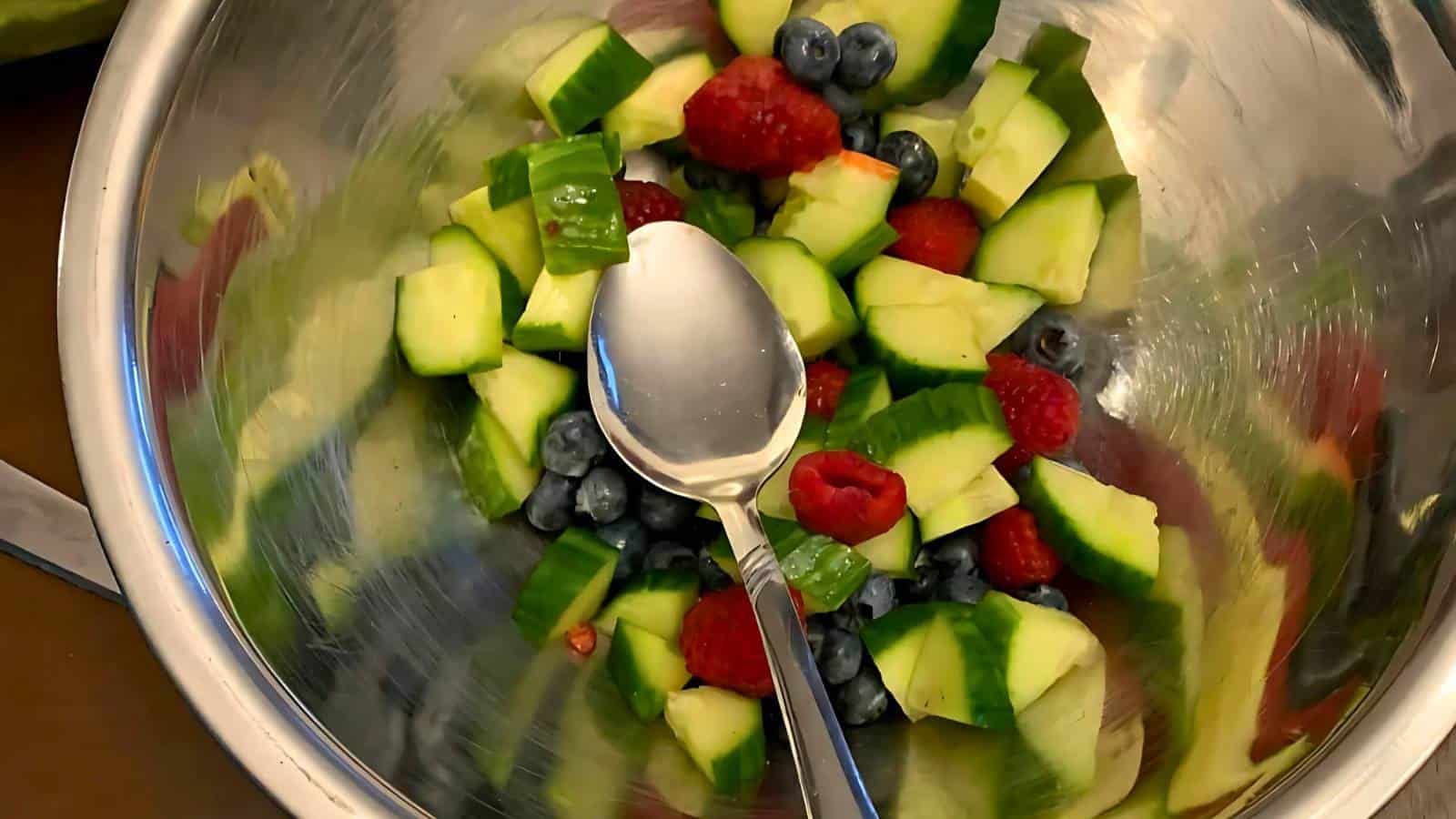 A metal bowl containing chopped cucumber, blueberries, raspberries, and a metal spoon.