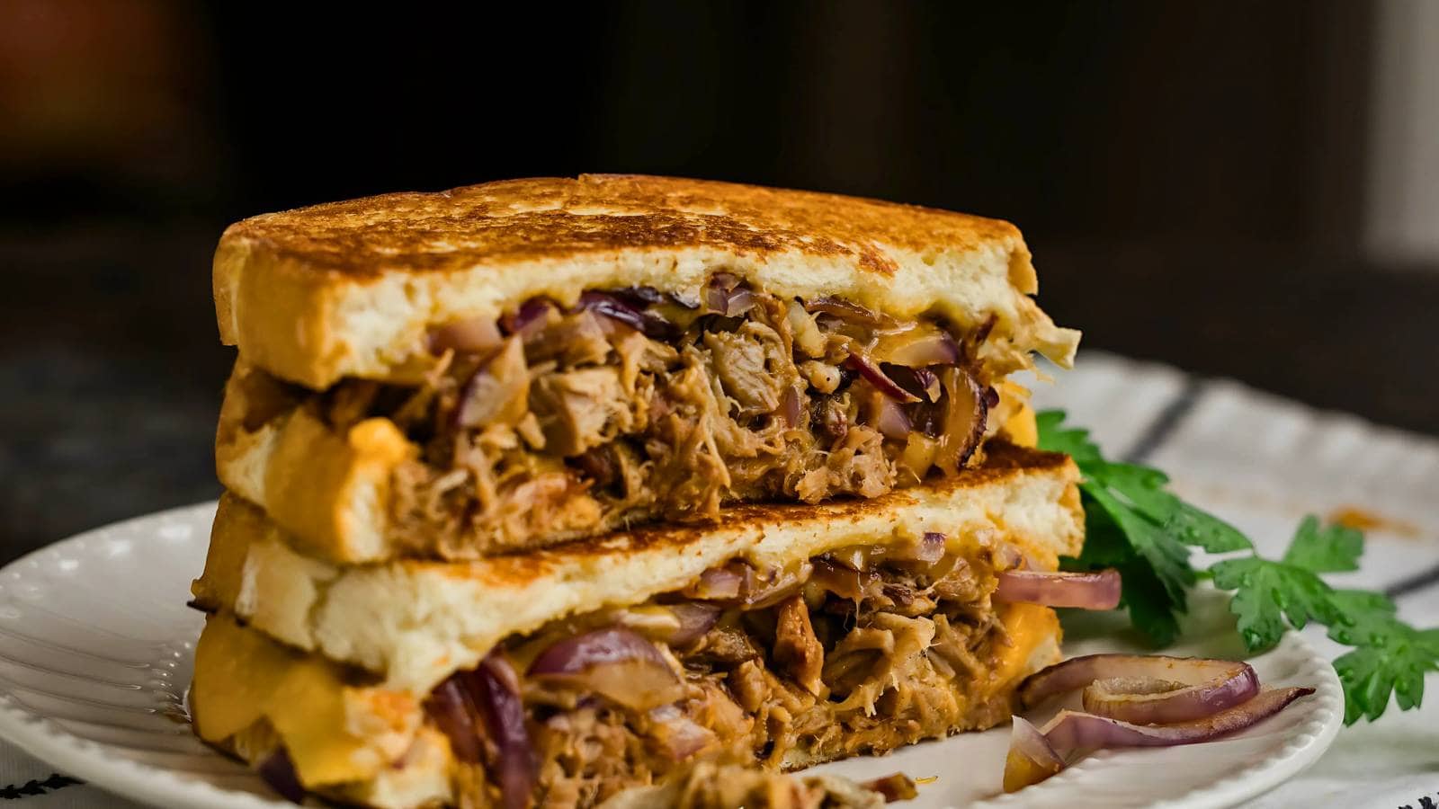 A grilled sandwich cut in half, filled with shredded meat, melted cheese, and sliced red onions, served on a white plate with a garnish of fresh herbs.