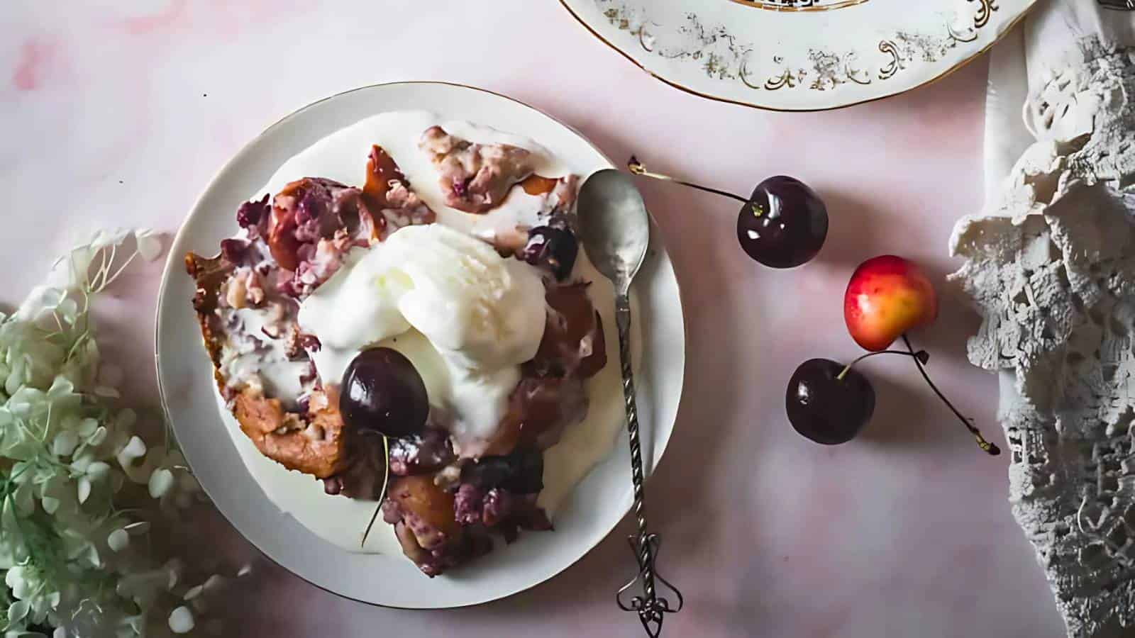 A plate with cherry dessert topped with ice cream and a cherry, a spoon, fresh cherries, a lace cloth, and part of a decorative plate on a pink surface.