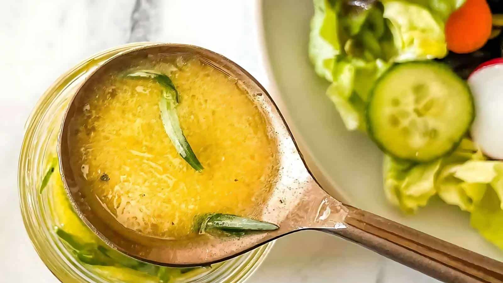 A ladle of vinaigrette with herbs is held above a glass bowl, next to a plate of salad with lettuce, cucumber, and radish.