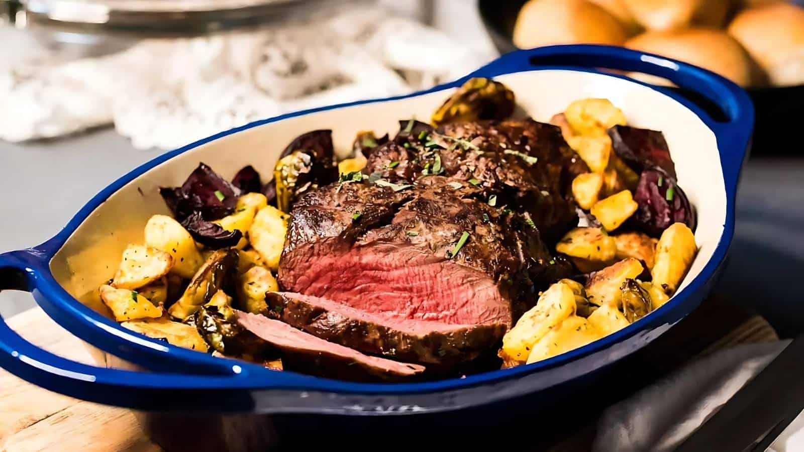 A blue baking dish contains sliced roast beef, cooked medium-rare, surrounded by roasted potatoes and vegetables.