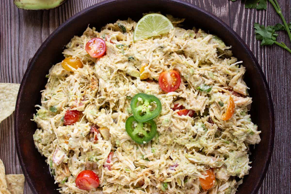 A bowl of Mexican chicken salad recipe features shredded chicken mixed with avocado, cherry tomatoes, jalapeño slices, and lime, served on a wooden table.