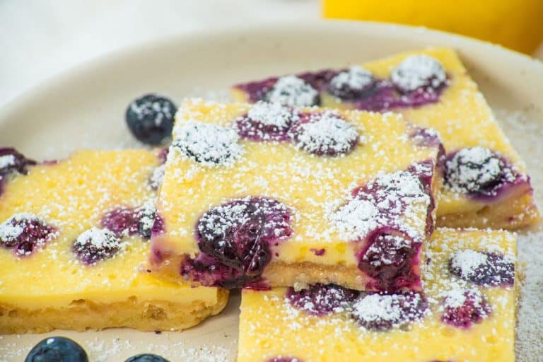 Three blueberry lemon bars topped with powdered sugar are arranged on a white plate, with a few fresh blueberries scattered nearby.