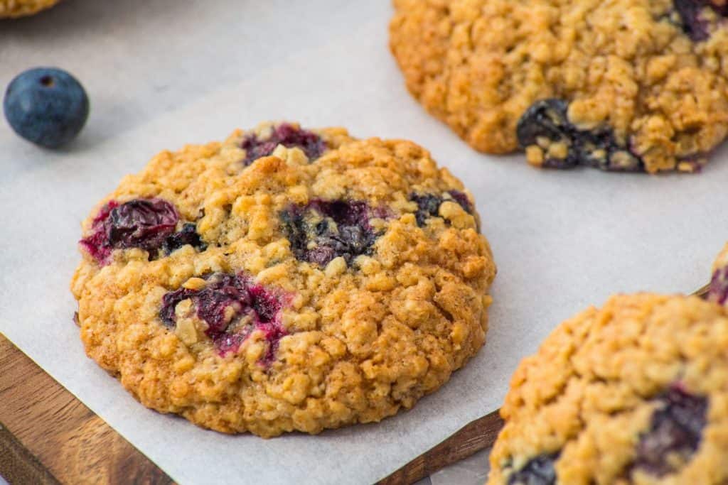 Oatmeal cookies with visible blueberries placed on parchment paper, with a single whole blueberry nearby.