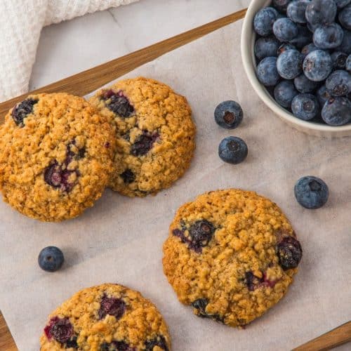 Five blueberry oatmeal cookies on parchment paper next to a bowl of fresh blueberries, placed on a wooden board.