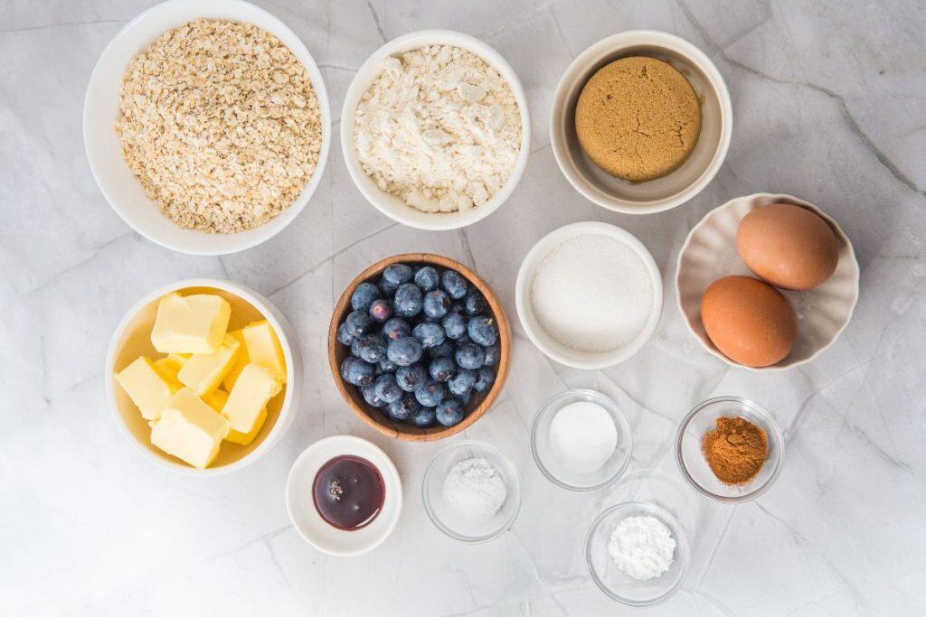 Bowls on a marble surface containing oats, flour, brown sugar, butter, blueberries, white sugar, eggs, jam, salt, baking powder, and cinnamon.