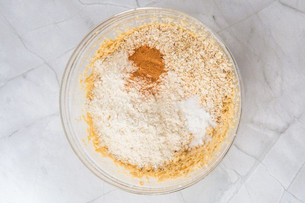 A glass bowl containing oats, white flour, cinnamon, baking soda, and a creamed mixture, placed on a marble countertop.