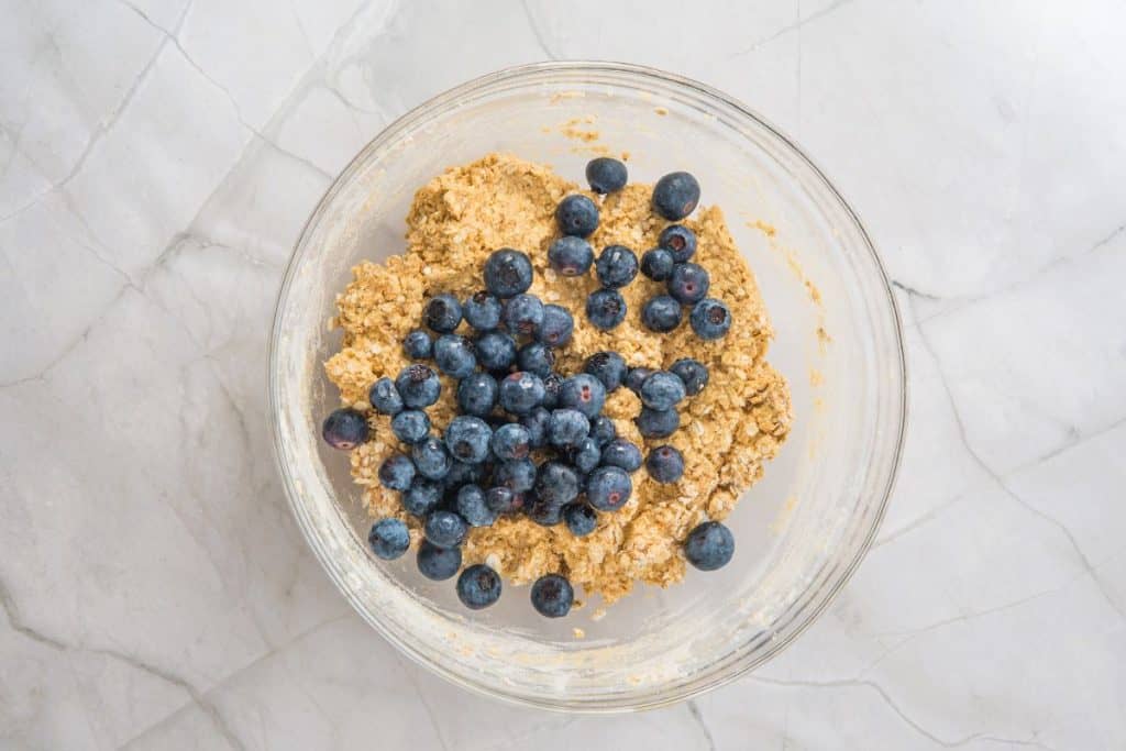 A glass bowl containing a mixture of dough and fresh blueberries sits on a white marble surface.