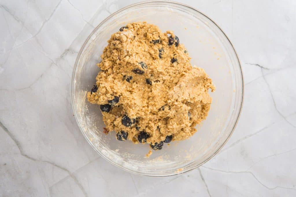 A glass bowl containing oatmeal cookie dough with visible raisins sits on a white marble surface.