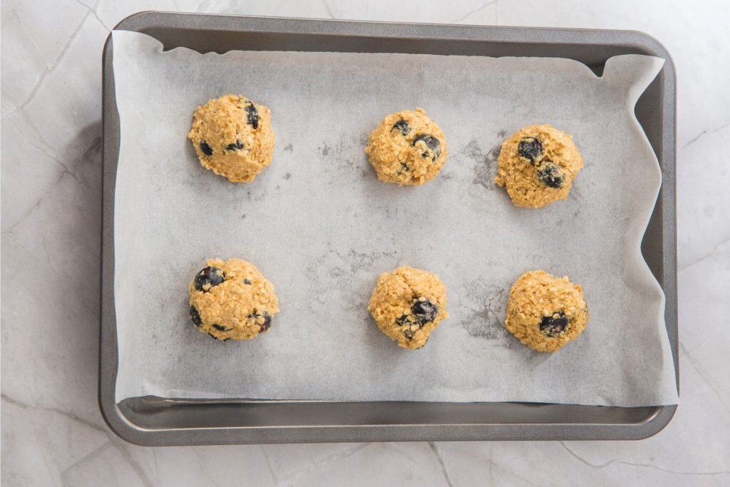 A metal baking tray lined with parchment paper holds six unbaked cookie dough balls with visible chocolate chips, spaced evenly apart.