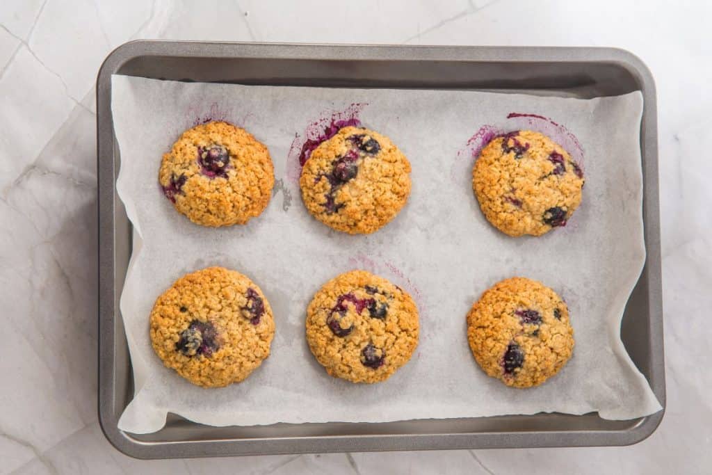 A baking tray with six blueberry oatmeal cookies on parchment paper, set on a light-colored marble surface.