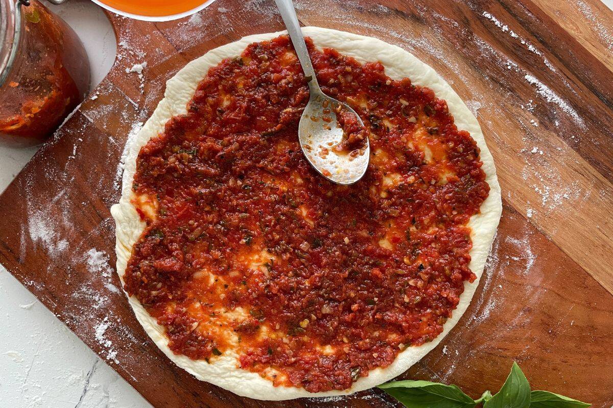 Unbaked pizza dough on a wooden board with zesty pizza sauce being spread on top using a spoon.