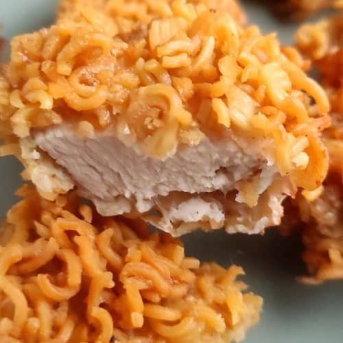 Close-up of a piece of chicken coated in crushed instant ramen noodles, with a visible white interior and crunchy exterior.