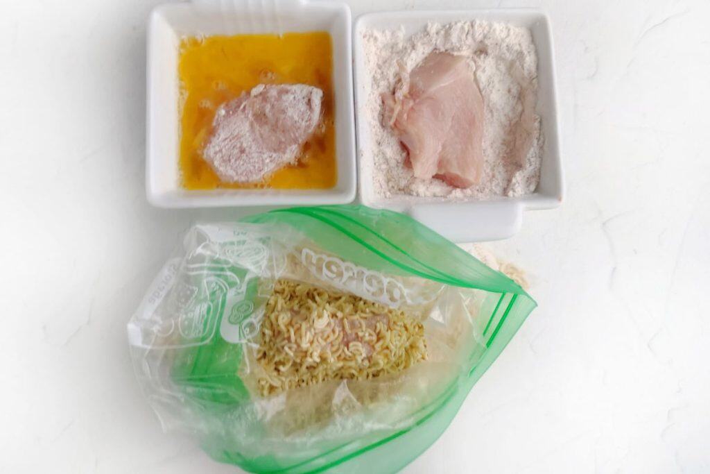 A piece of raw chicken is coated in flour, dipped in egg wash, and placed in a plastic bag with crushed instant noodles for breading.