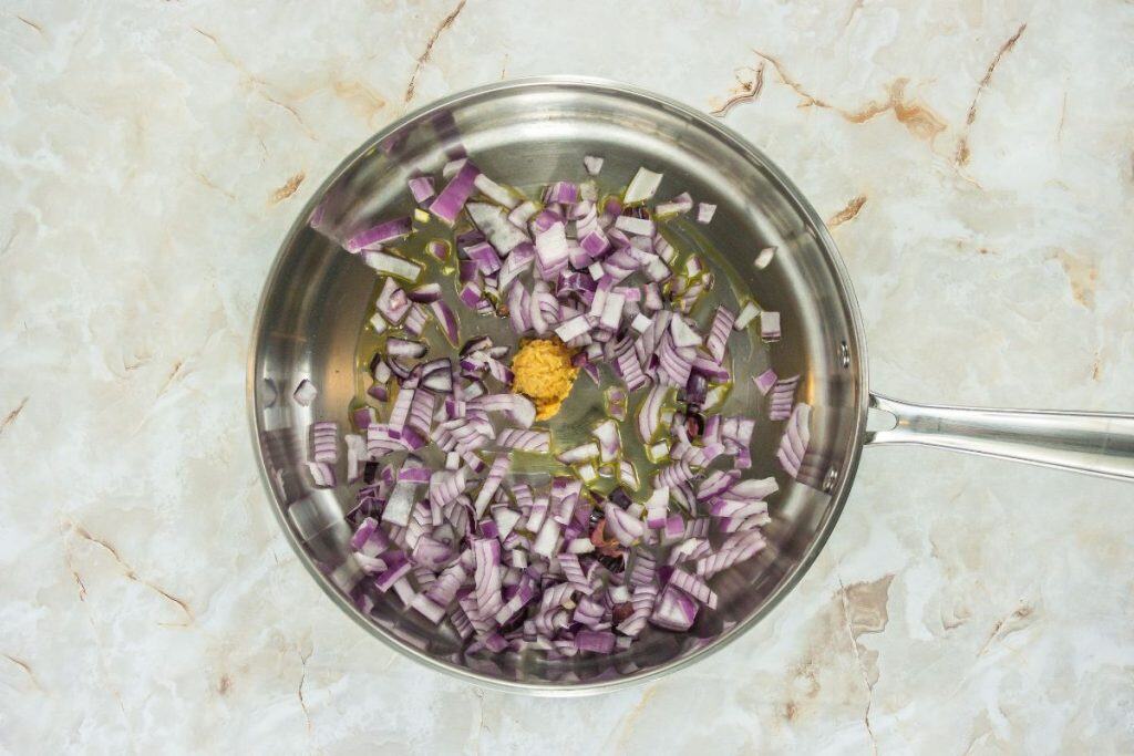 Chopped red onions and a small amount of minced garlic or ginger in a stainless steel pan on a light marble surface.