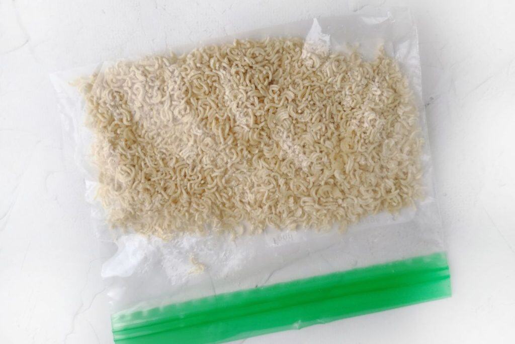 Uncooked instant ramen noodles inside a resealable plastic bag with a green zipper on a white surface.