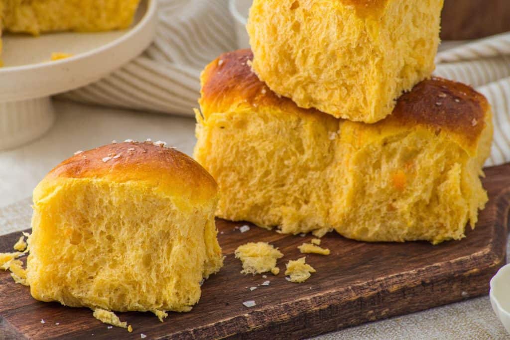 Three soft, golden sweet potato dinner rolls are stacked on a wooden board with crumbs scattered around, and a fourth roll sits in front of the stack.
