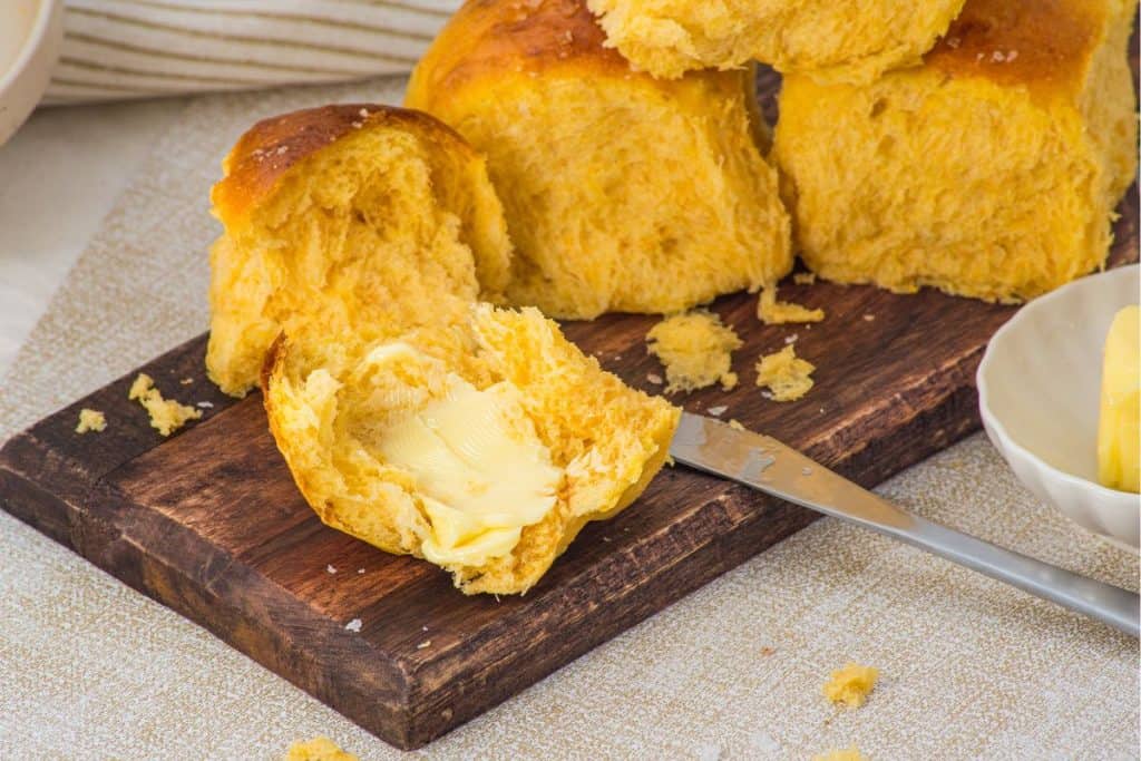 A piece of golden brown bread roll with a pat of butter, sliced open on a wooden board, surrounded by more rolls and a butter knife.