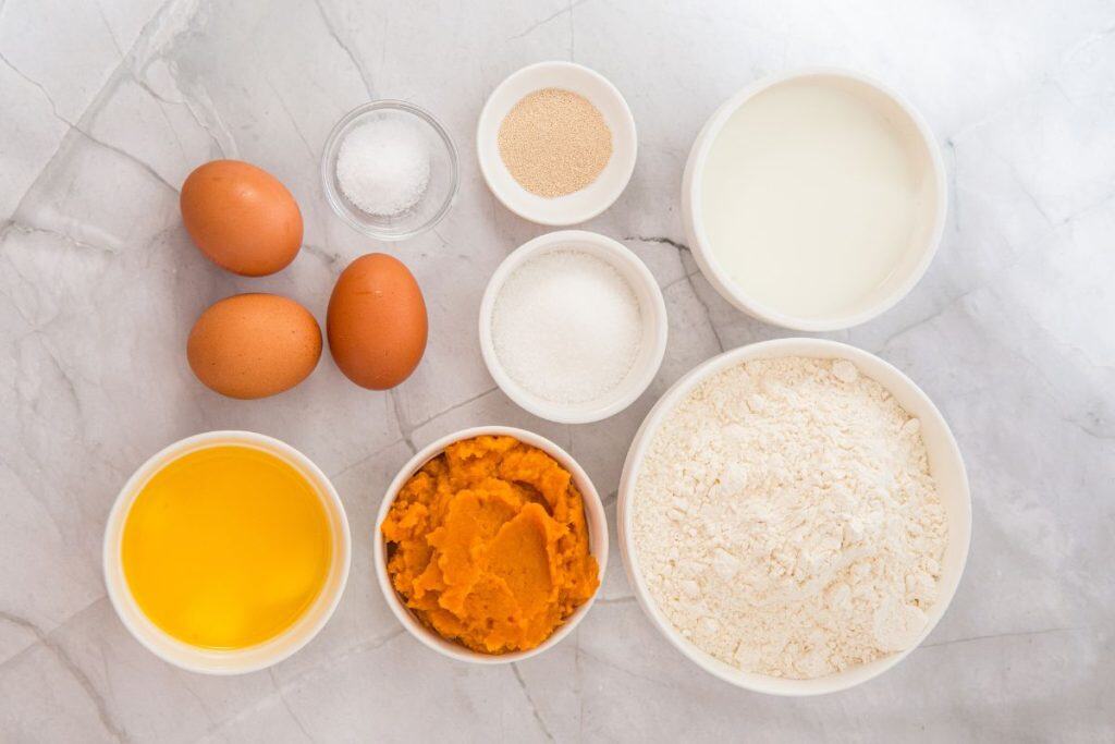 Ingredients for baking sweet potato dinner rolls on a marble surface: eggs, salt, yeast, milk, butter, sugar, flour, and pumpkin puree in separate bowls.