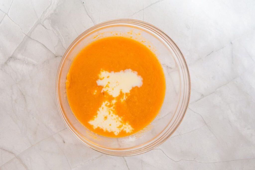 A glass bowl containing orange puree with a small amount of white liquid on top, placed on a light marble surface.