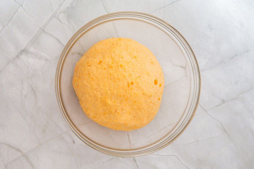 A round ball of orange dough sits in a clear glass bowl on a light gray marble surface.