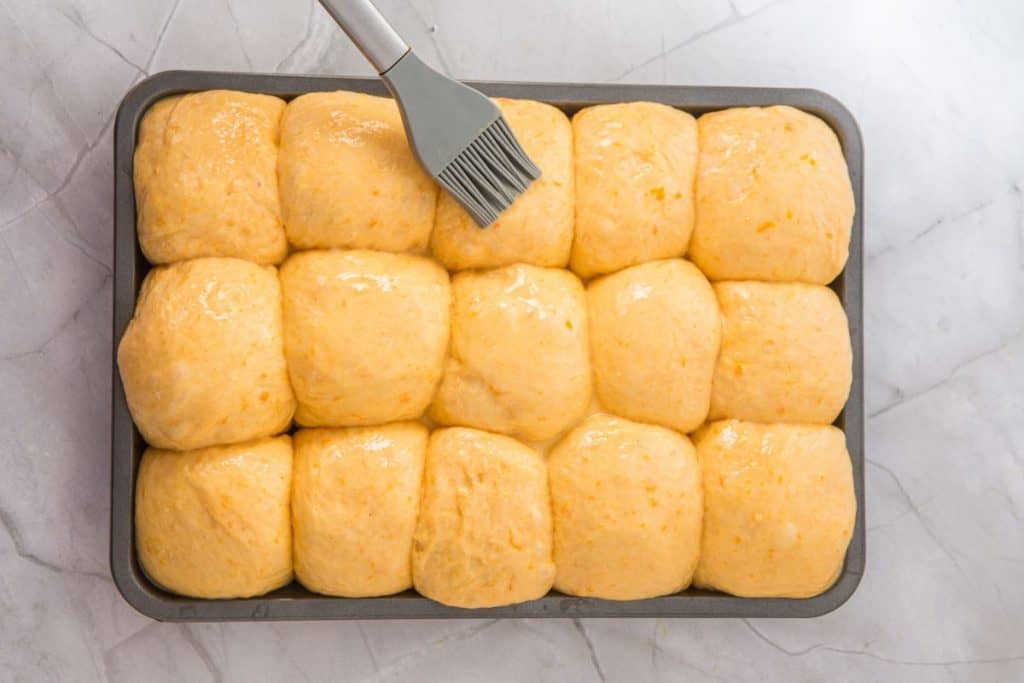 A tray of sixteen unbaked dinner rolls is being brushed with an egg wash using a silicone pastry brush on a marble countertop.