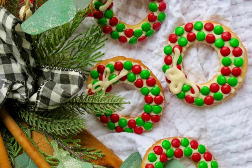 Four cookies decorated as Christmas wreaths with red and green candies and white icing bows are placed on a white cloth next to greenery and a black-and-white checkered ribbon for a Christmas cookie exchange.