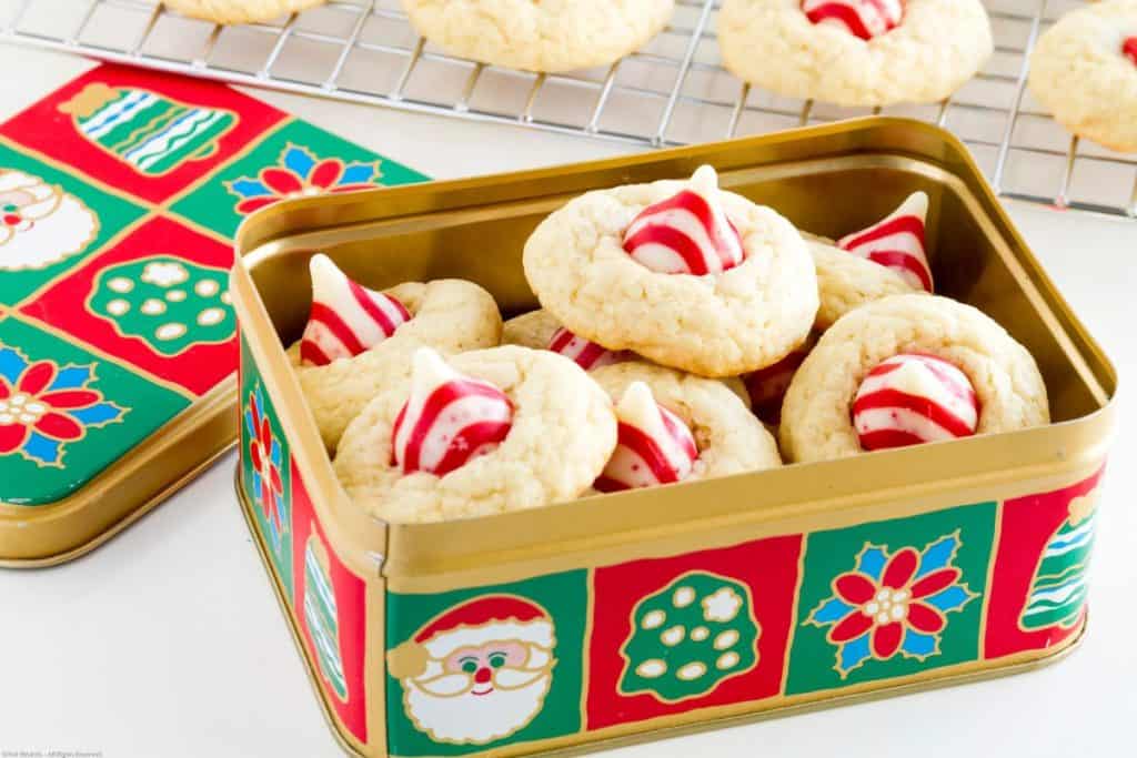 A festive tin filled with sugar cookies topped with red and white striped chocolate kisses, with more cookies cooling on a rack in the background.