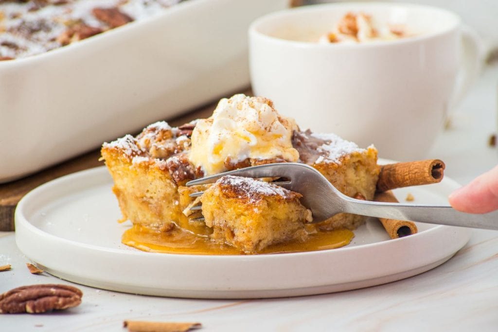 A fork cutting into a slice of eggnog french toast casserole topped with ice cream and powdered sugar, served on a white plate with syrup and cinnamon sticks.