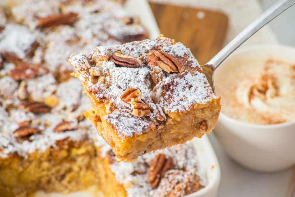 A close-up of a slice of eggnog French toast casserole topped with powdered sugar and pecans, held above a baking dish with more bread pudding and a cup of coffee in the background.