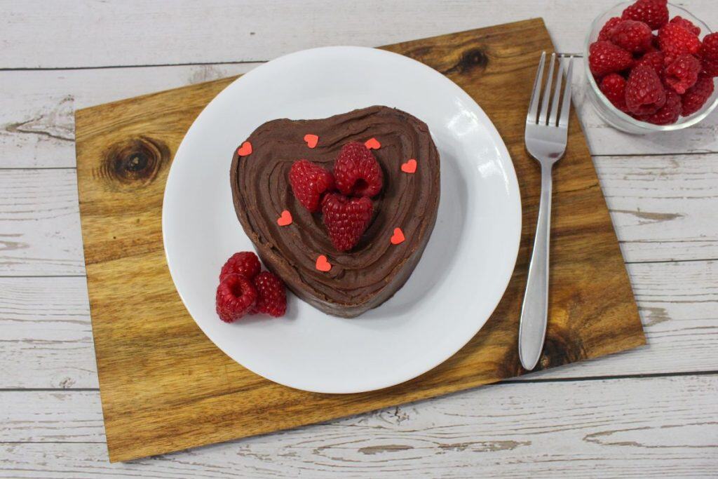A heart-shaped chocolate cake with raspberries and red heart sprinkles on a white plate, placed on a wooden board with a fork and a bowl of raspberries nearby.