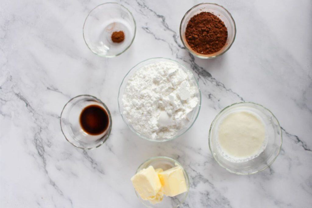 Six glass bowls on a marble surface containing cocoa powder, powdered sugar, butter, vanilla extract, milk, and a mix of salt and cinnamon, ingredients for chocolate Valentine's Day cake.