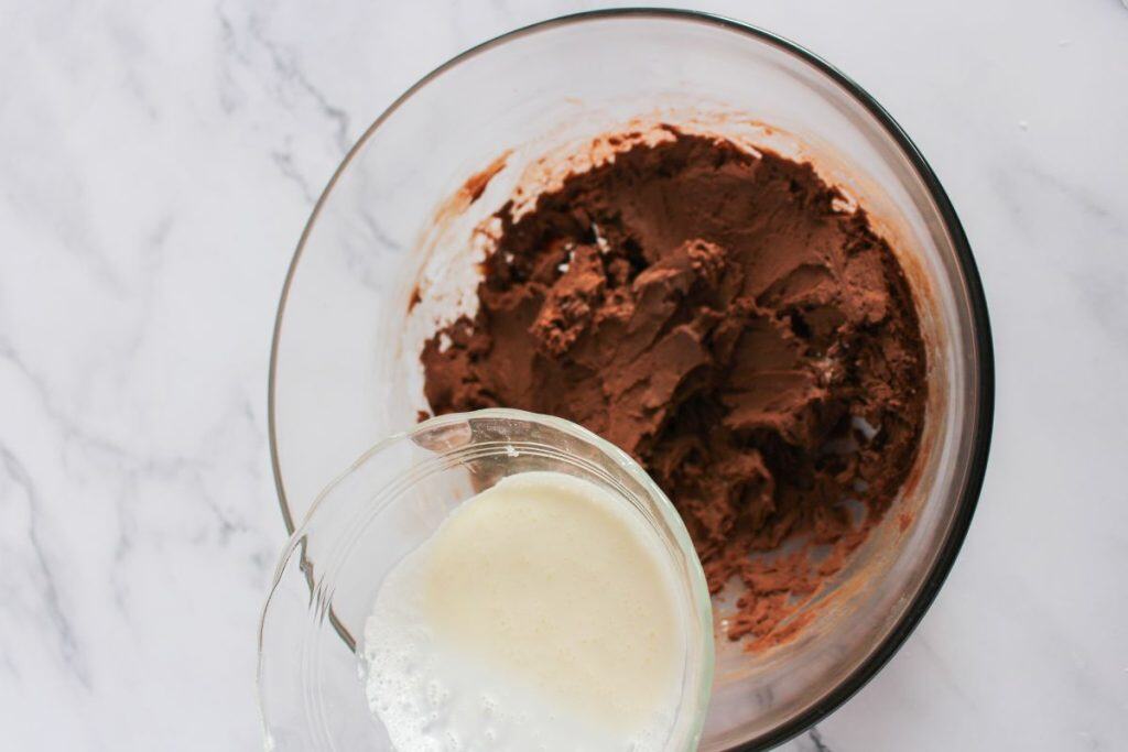 A glass bowl with chocolate batter and another bowl pouring cream into it, on a white marble surface.