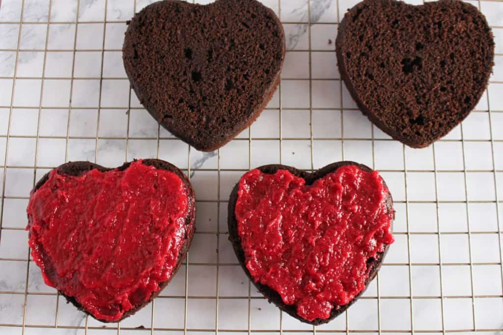 Four heart-shaped chocolate cake layers on a cooling rack; two are plain, and two are topped with a layer of red fruit jam or filling.