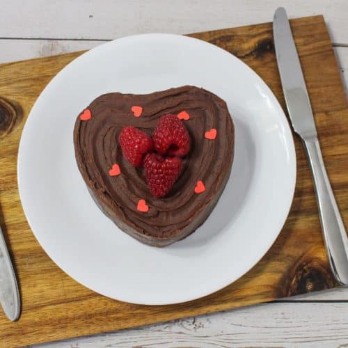 A heart-shaped chocolate cake with raspberries and red heart sprinkles on a white plate, set with a fork and knife on a wooden board; a bowl of raspberries is beside it.