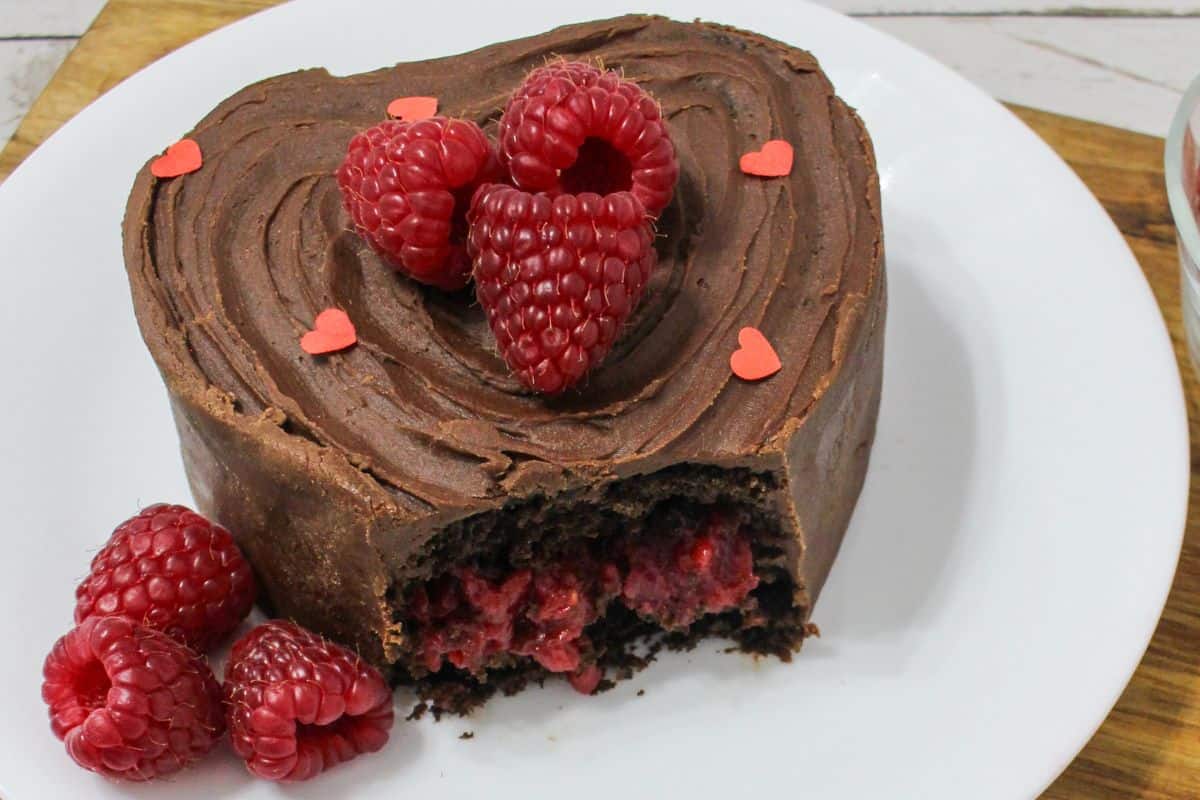 A heart-shaped chocolate cake with a bite taken out, filled with raspberries, topped with raspberries and small red heart sprinkles, on a white plate.