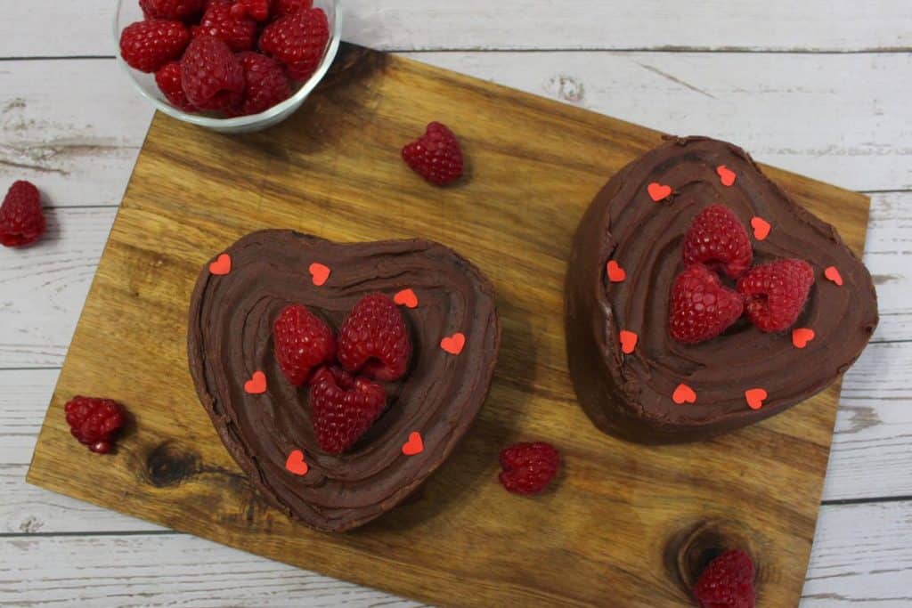 Two heart-shaped chocolate cakes topped with raspberries and small red heart decorations on a wooden board, with a bowl of raspberries nearby.