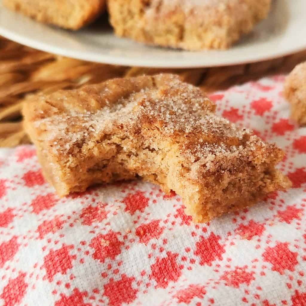 A piece of baked churro bar with a bite taken out, resting on a red and white patterned cloth, with more baked churro bars on a plate in the background.